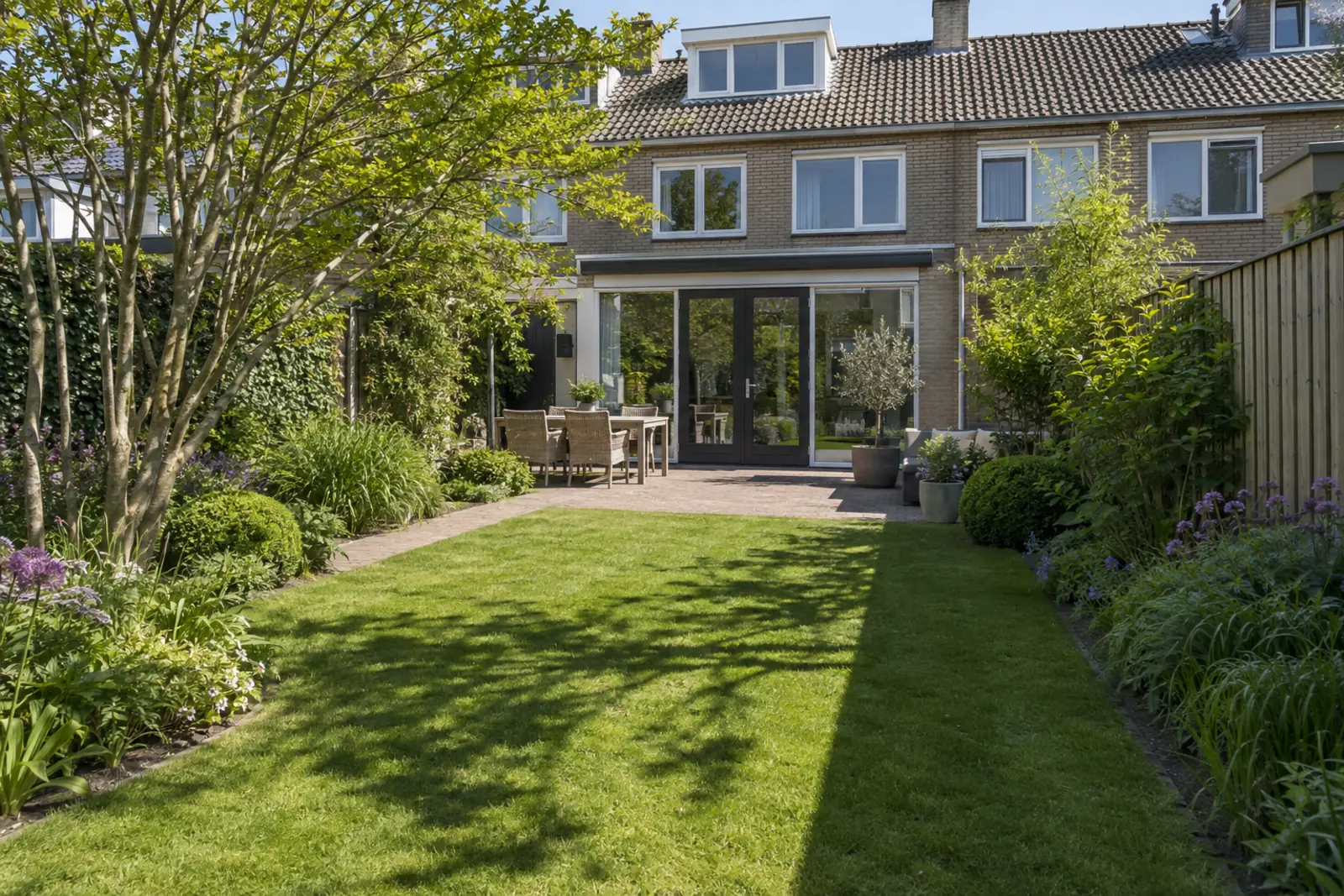 Achtertuin op een zonnige dag, gezien vanaf achterin: scherpe schaduwen van schutting en boom over border en gazon, het terras bij het huis half in de zon