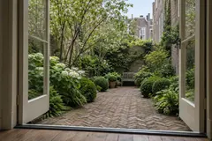 Kleine ommuurde Amsterdamse stadstuin door openstaande tuindeuren: klinkers in visgraat, buxusbollen, witte hortensia's, een meerstammige boom en een bankje