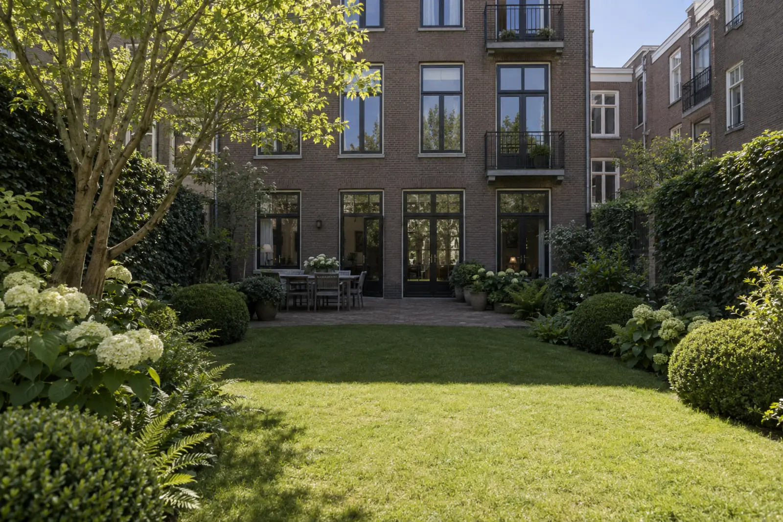 Smalle stadstuin ten noorden van een hoog bakstenen woonblok, gezien vanaf het einde van de tuin. De schaduw van het blok bedekt het terras en de eerste helft van het gazon, het deel vooraan ligt in de zon.