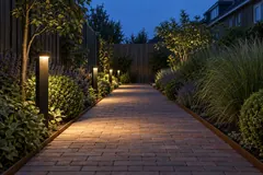 Een klinkerpad tussen twee borders in de avond, laag van de grond gezien, met slanke zwarte staande lampen die elk een plas warm licht op de stenen leggen